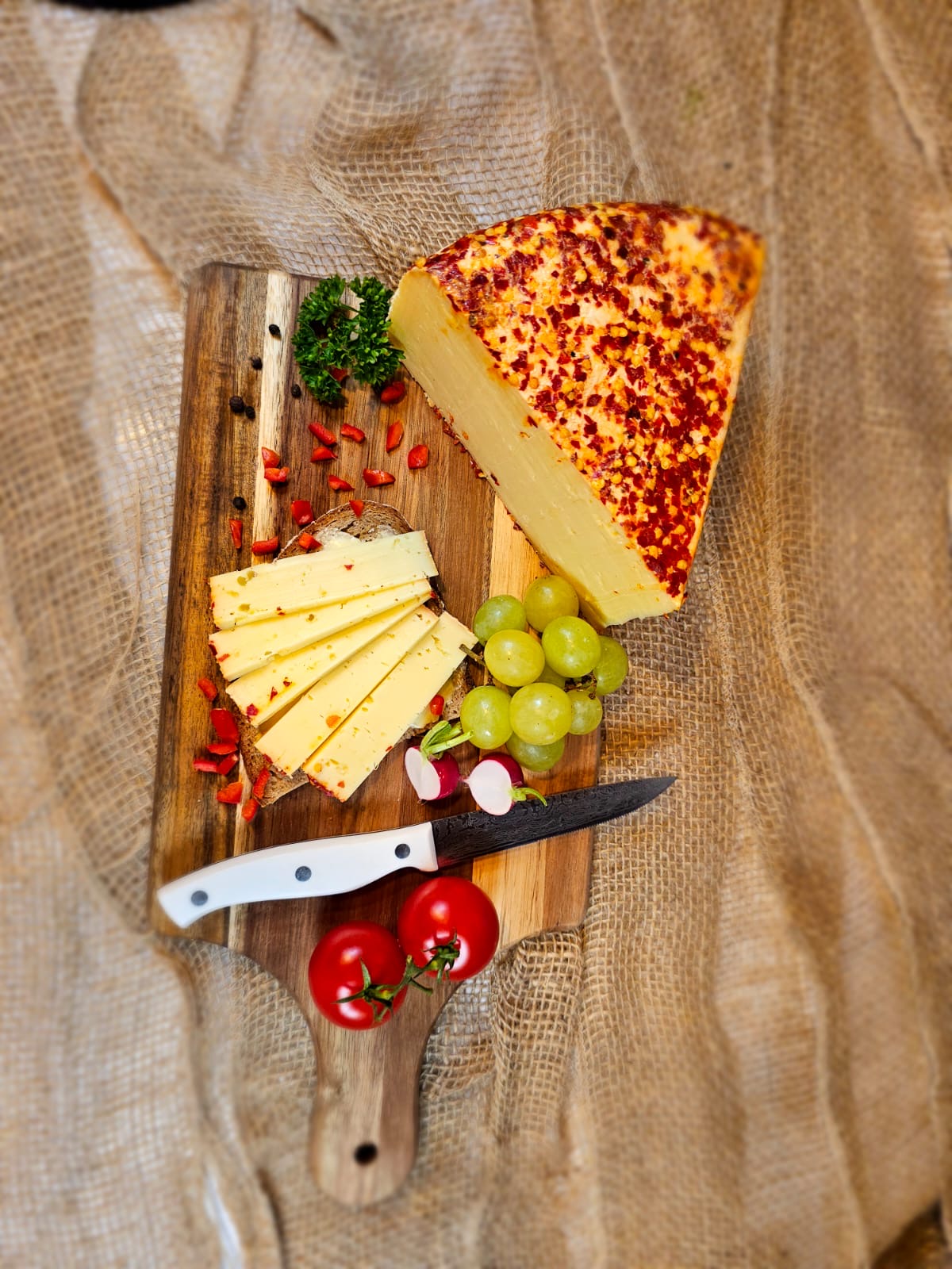 Bergkäse auf einem Holzbrett mit Chili verfeinert Chilibeer Käse mit roten Tomaten, einem Messer und einzelnen Scheiben. Der Hintergrund besteht aus Jute Stoff